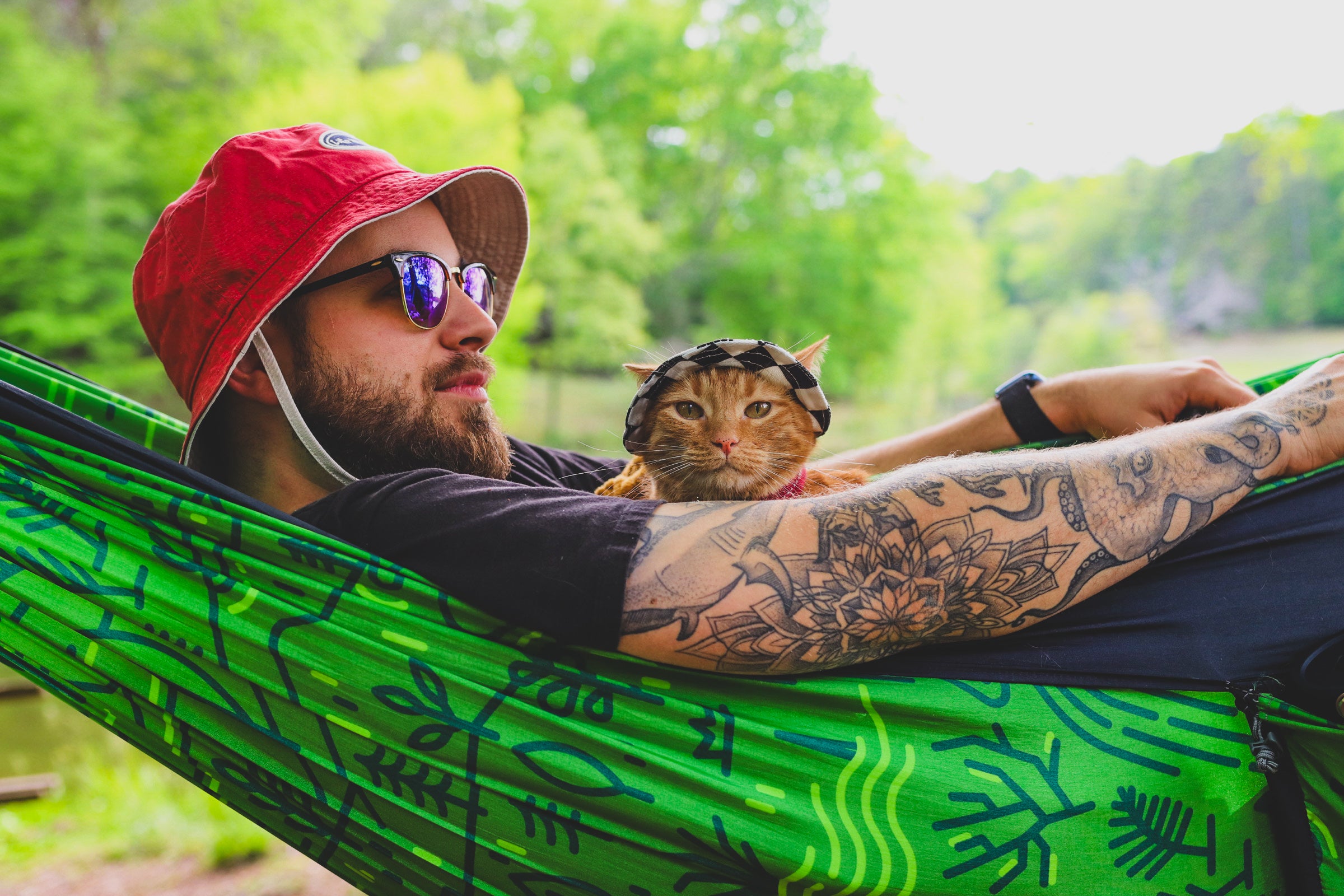Person relaxing in a hammock outdoors with an orange cat wearing a small hat nestled beside them.